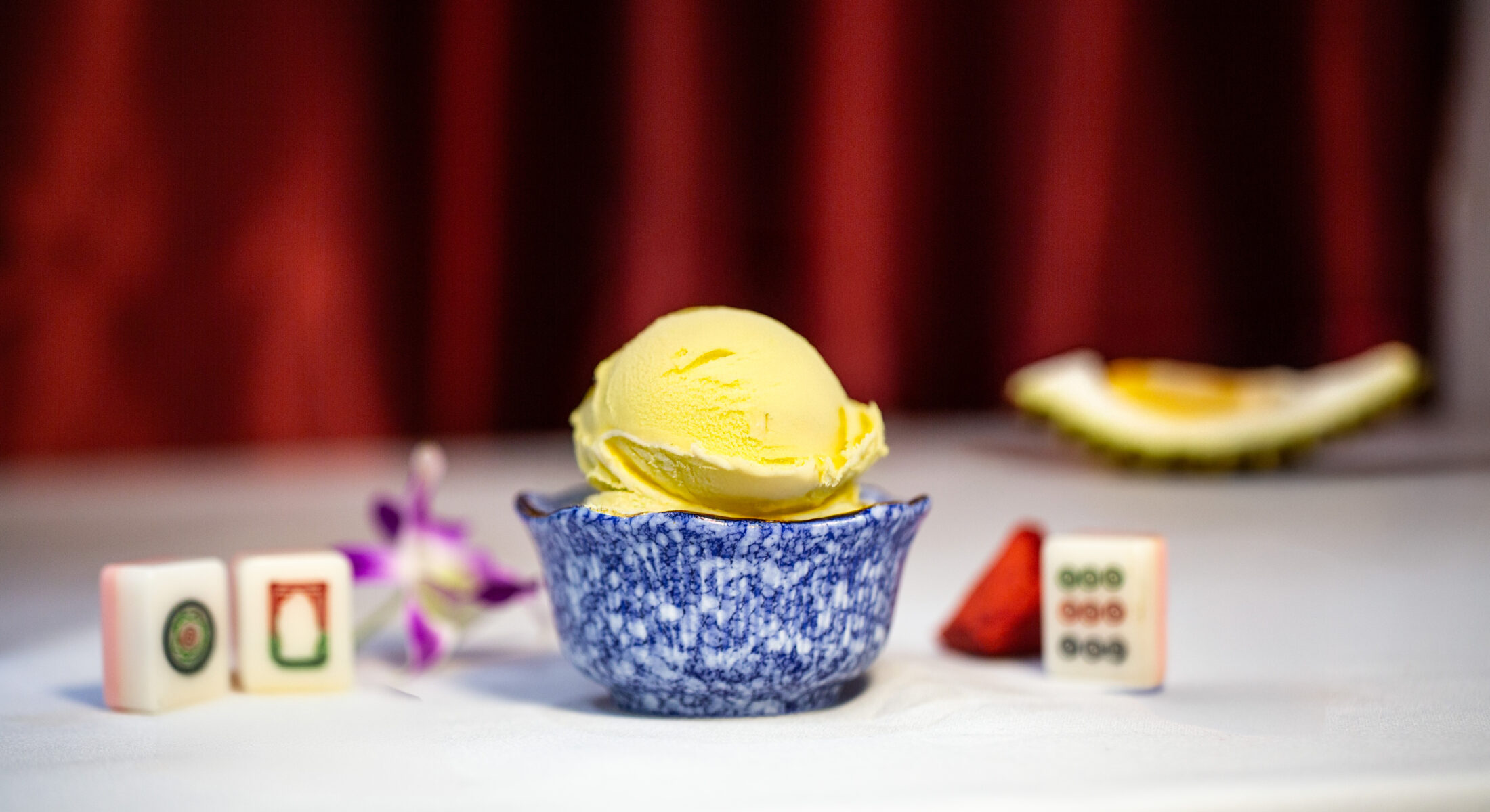 A scoop of creamy D24 durian ice cream in a blue marble bowl, with mahjong tiles and a durian wedge in the background.