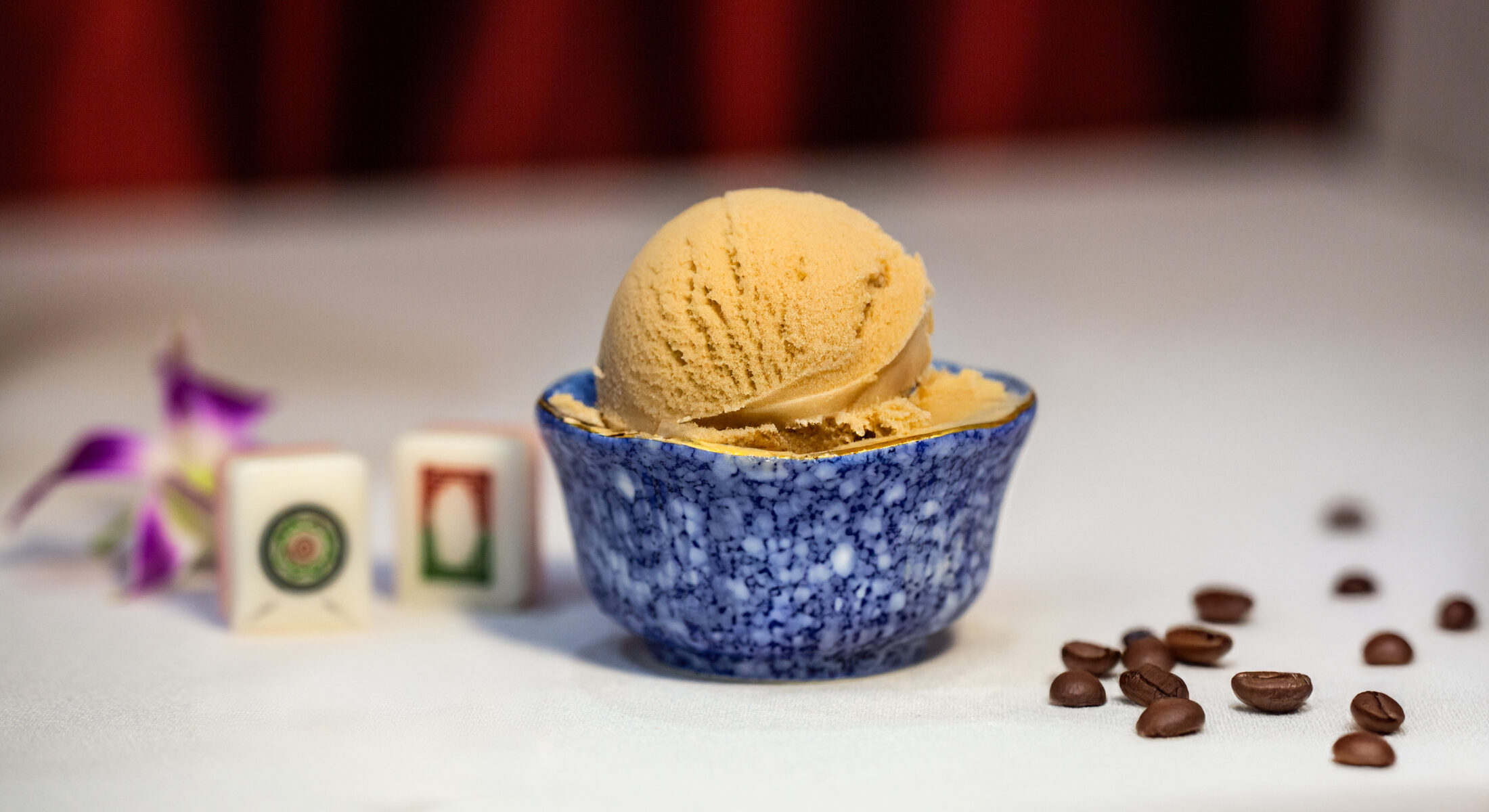 A scoop of coffee-hued Kopi Peng ice cream in a blue marble bowl, with scattered coffee beans and mahjong tiles.