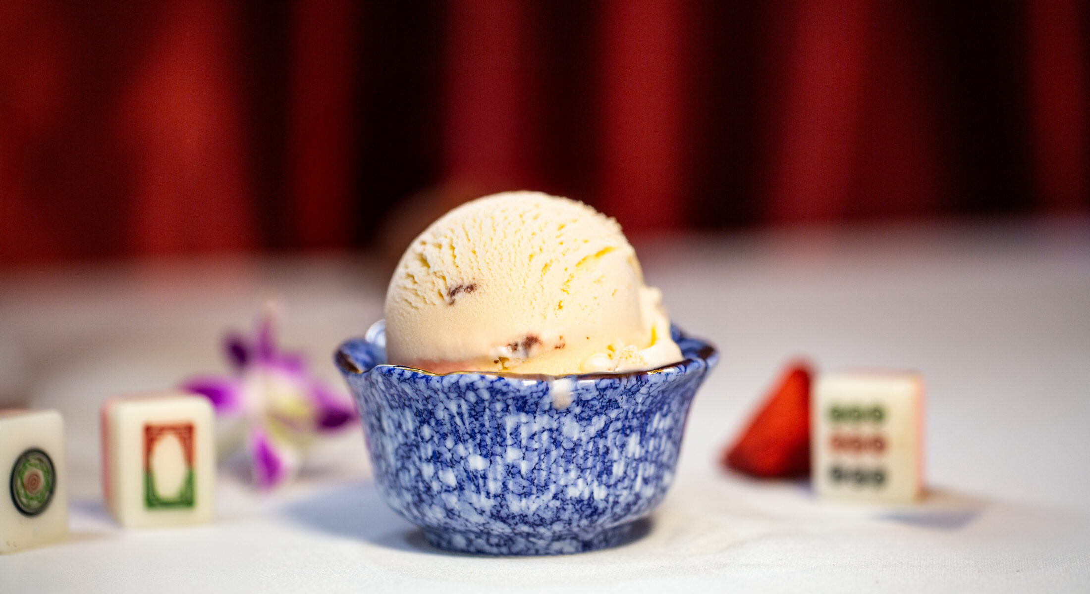 A scoop of Mexican vanilla ice cream in a blue marble bowl, with mahjong tiles and a strawberry garnish in the background.