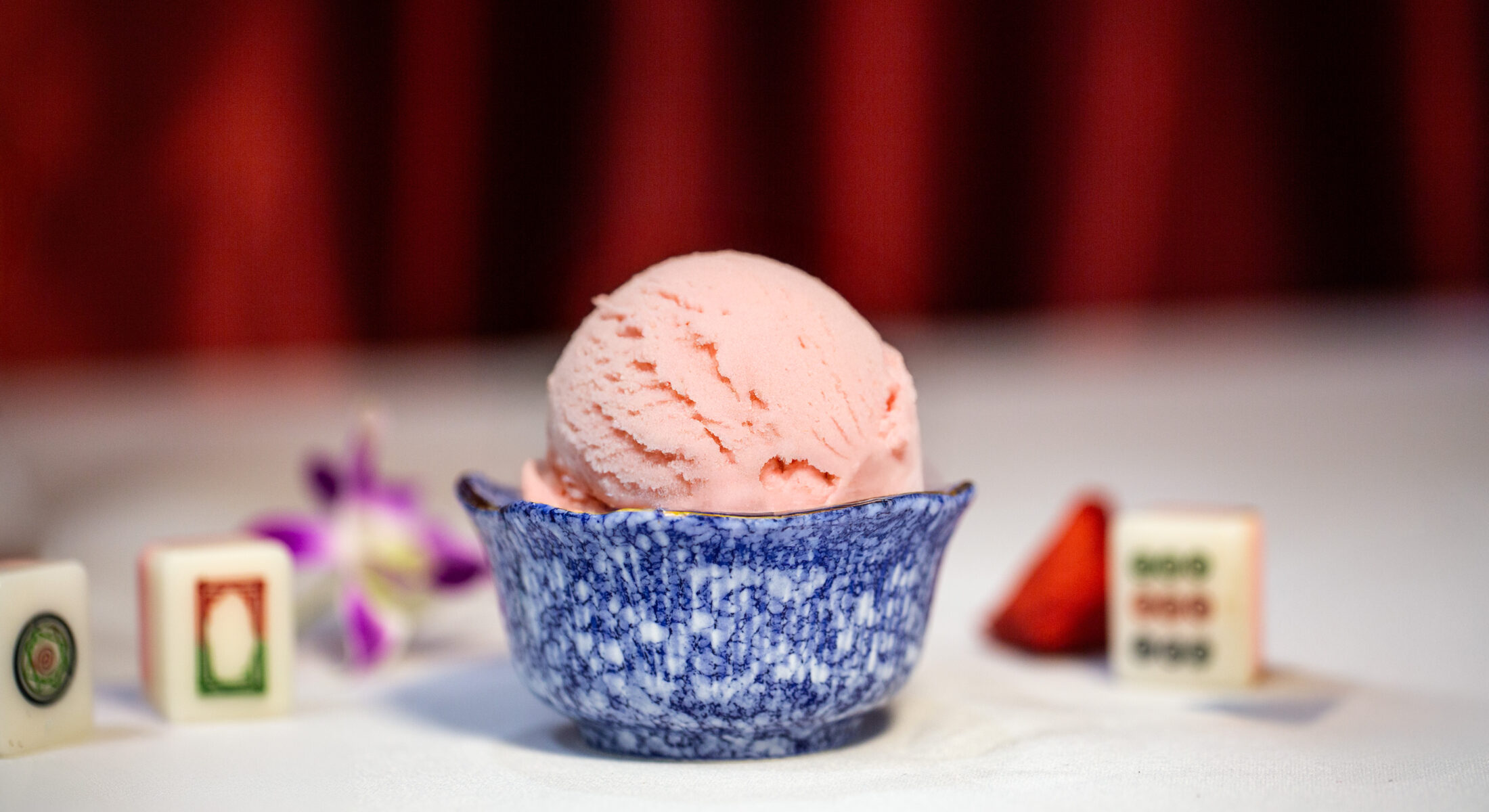 A scoop of pink watermelon sorbet in a blue marble bowl, with mahjong tiles and a strawberry garnish.