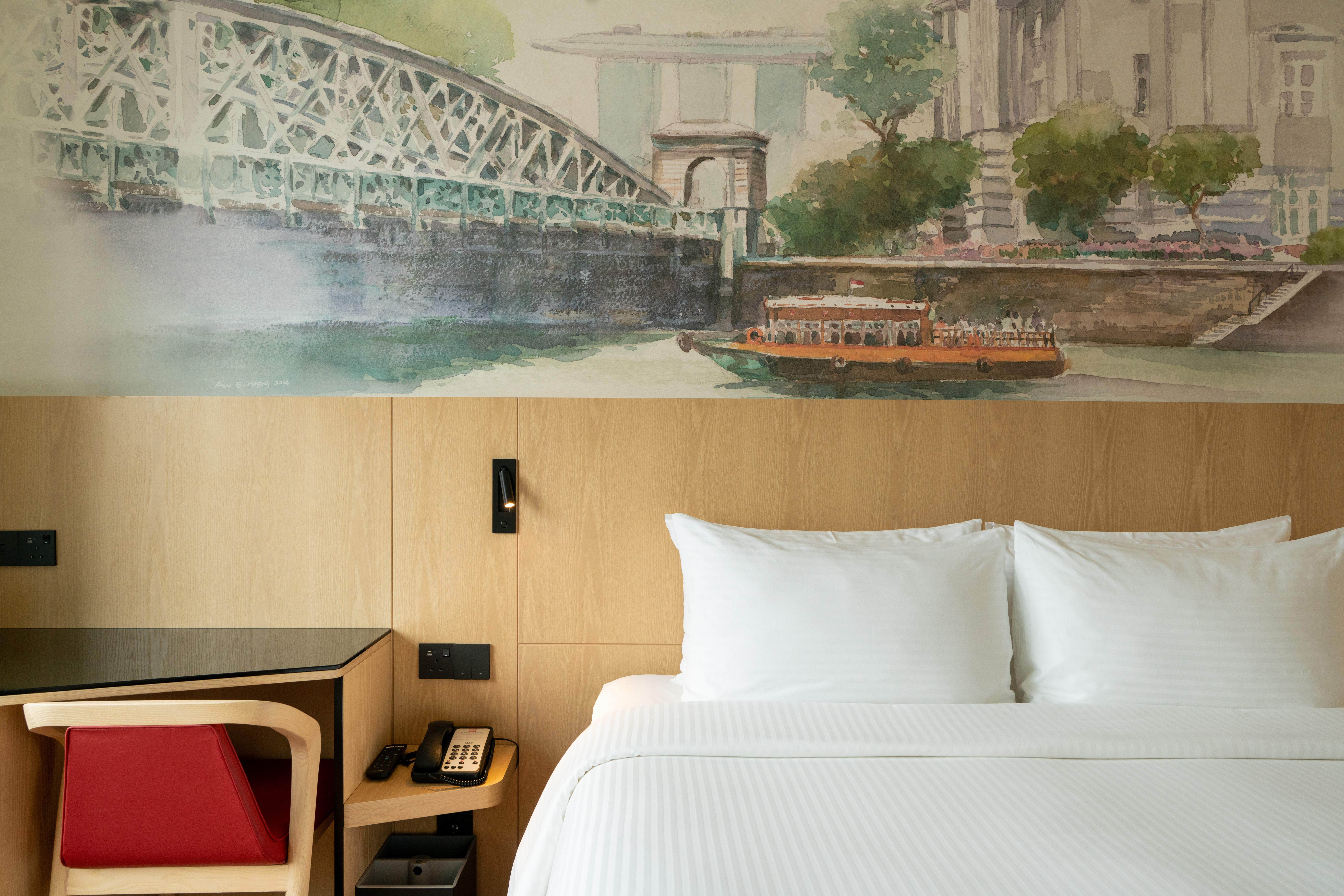 Spacious hotel room featuring a comfortable bed with a decorative headboard mural illustrating the Singapore River, a study table, and a bay window offering natural light.