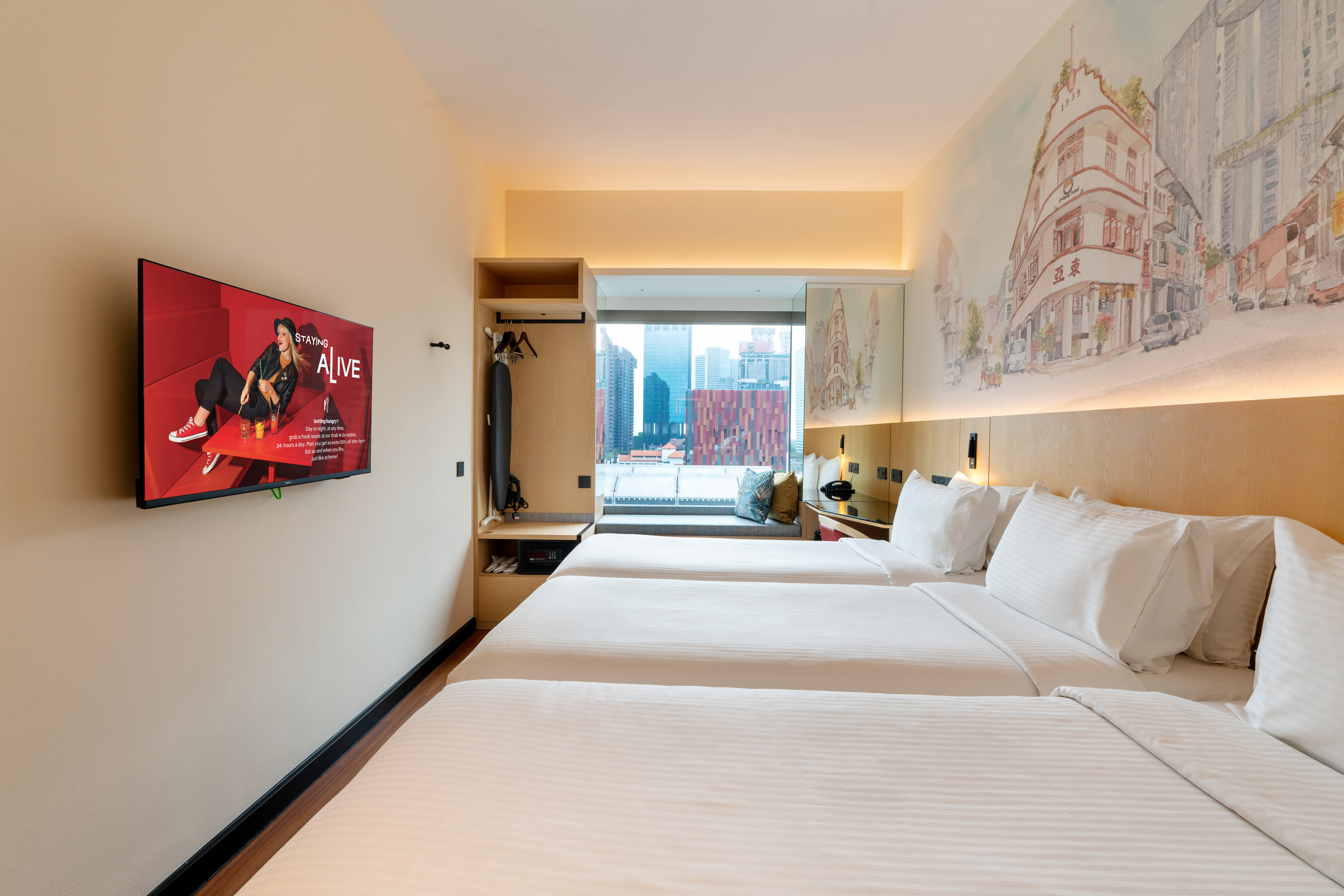 Hotel room featuring Triple comfortable beds with a decorative headboard mural illustrating a Singapore street with public housing at the background, a study table, and a bay window offering natural light.
