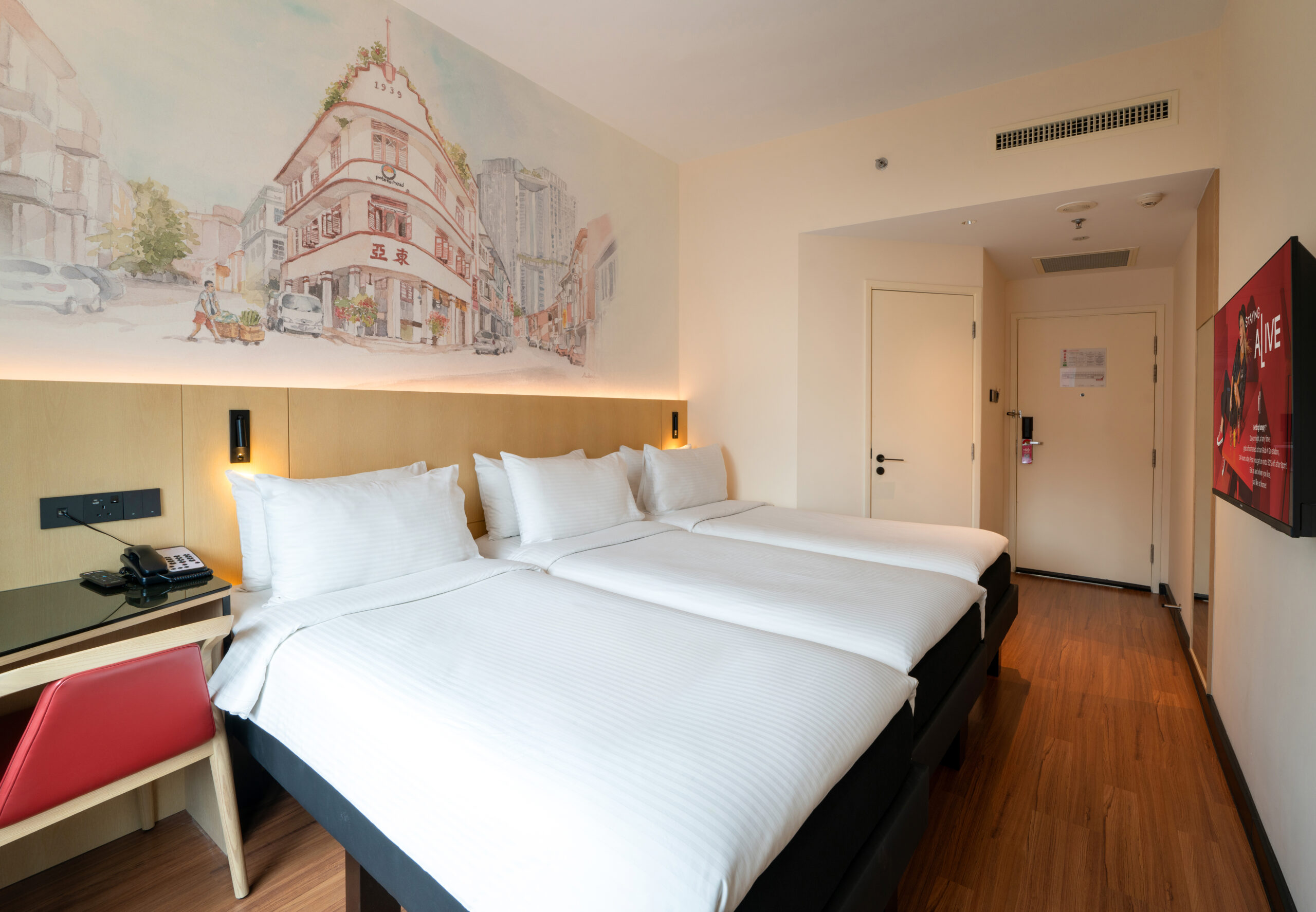 Hotel room featuring Triple comfortable beds with a decorative headboard mural illustrating a Singapore street with public housing at the background, a study table, and a bay window offering natural light.