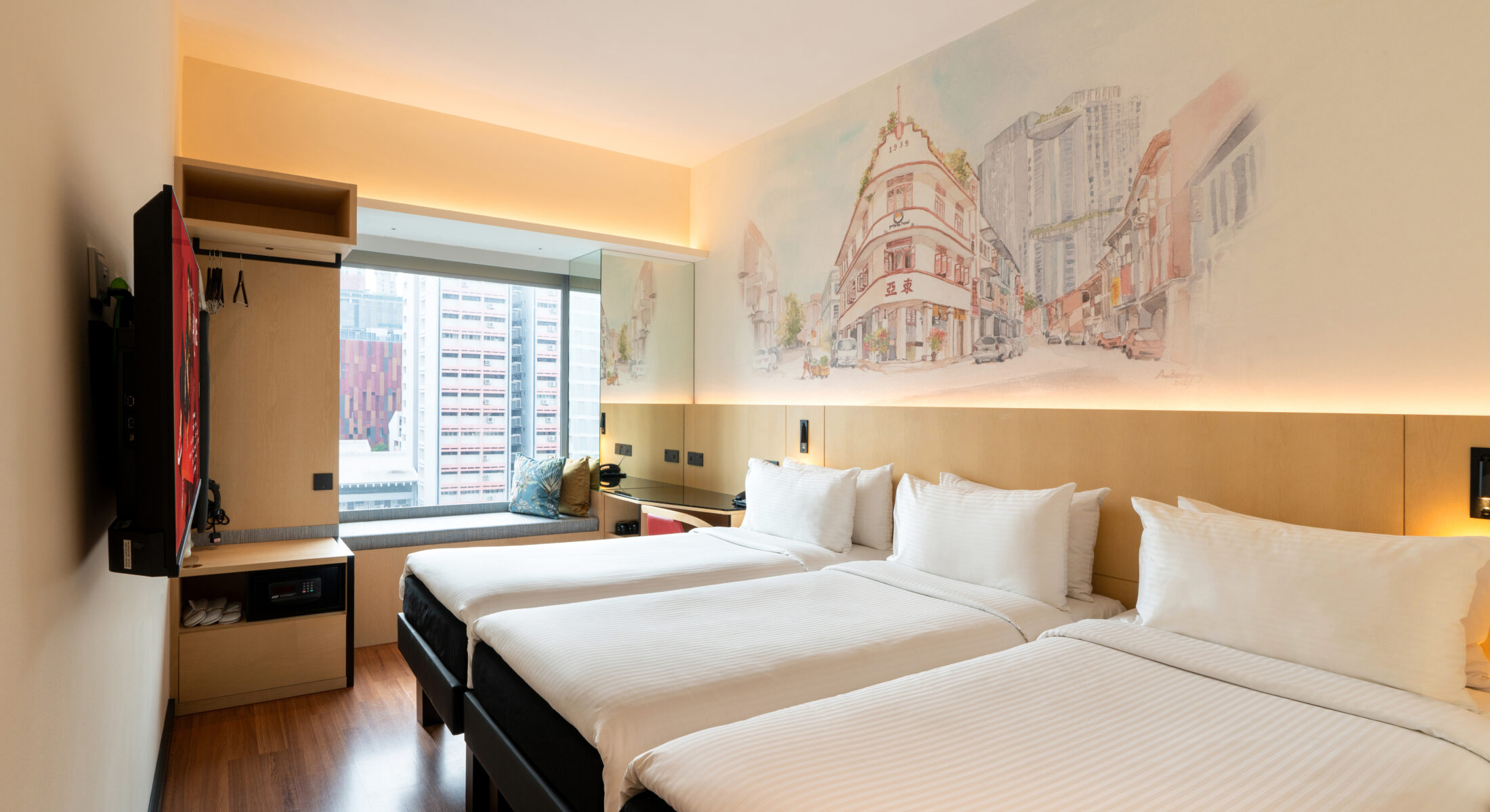 Hotel room featuring Triple comfortable three single beds with a decorative headboard mural illustrating a Singapore street with public housing at the background, a study table, and a bay window offering natural light.