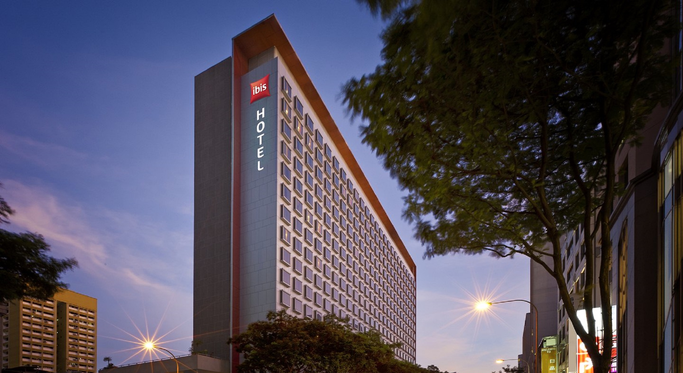 Exterior view of ibis Singapore on Bencoolen hotel building at dusk, with illuminated signage and city street.