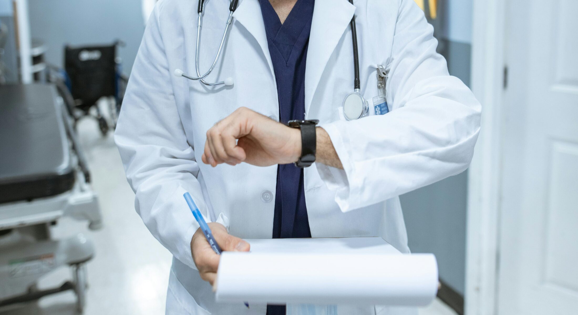 Healthcare professional in a white coat reviewing notes in a hospital corridor with medical equipment in the background