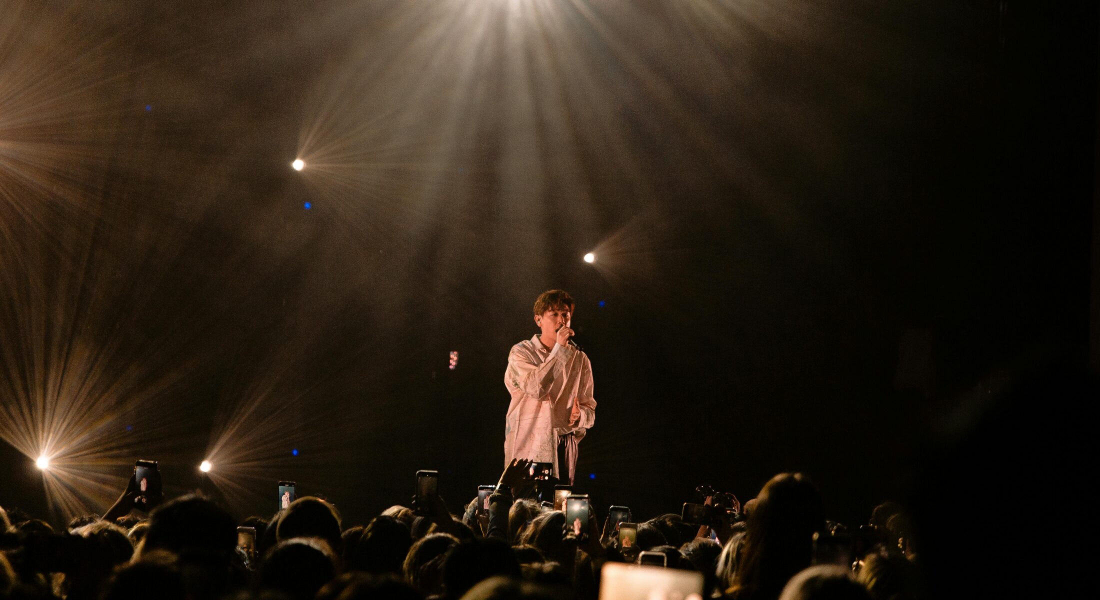 Singer performing live on stage at a concert with warm stage lighting and an audience capturing the moment on their phones in Singapore