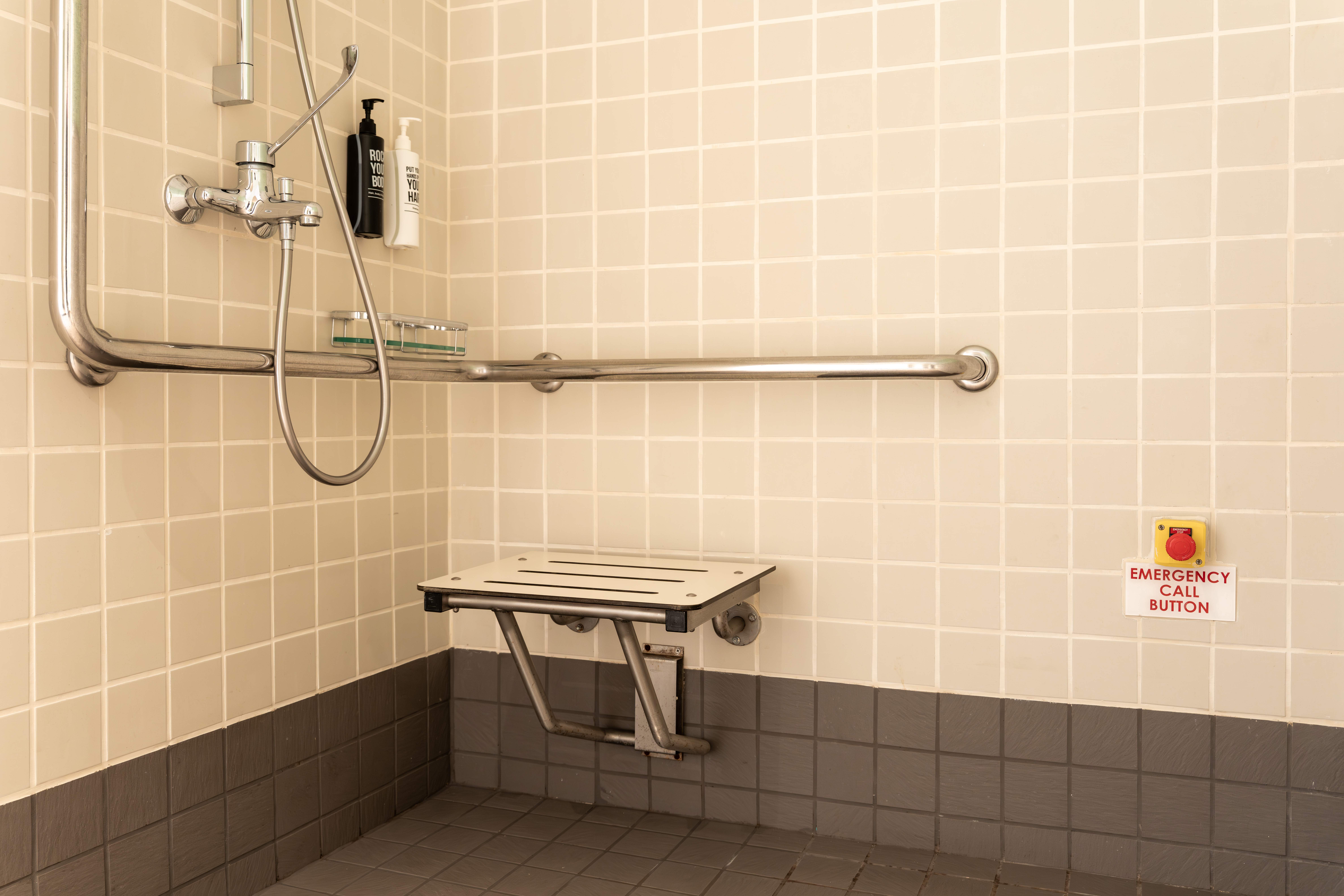 Close-up of accessible roll-in shower at ibis Singapore on Bencoolen featuring support grab bars, foldable shower seat, handheld shower, and emergency assistance button.
