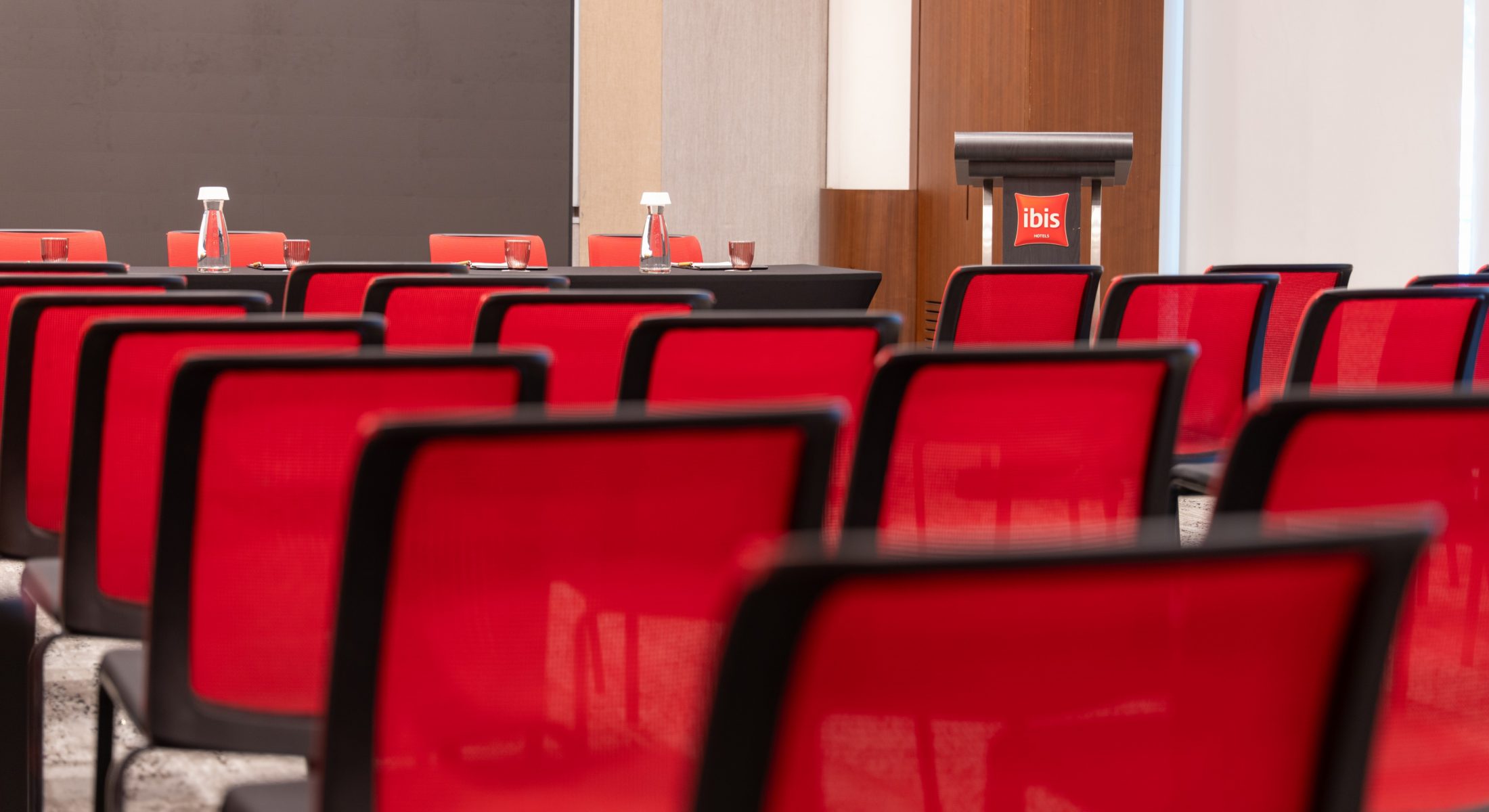 Rows of red meeting chairs facing a panel table with water bottles and glasses, beside an ibis-branded lectern.