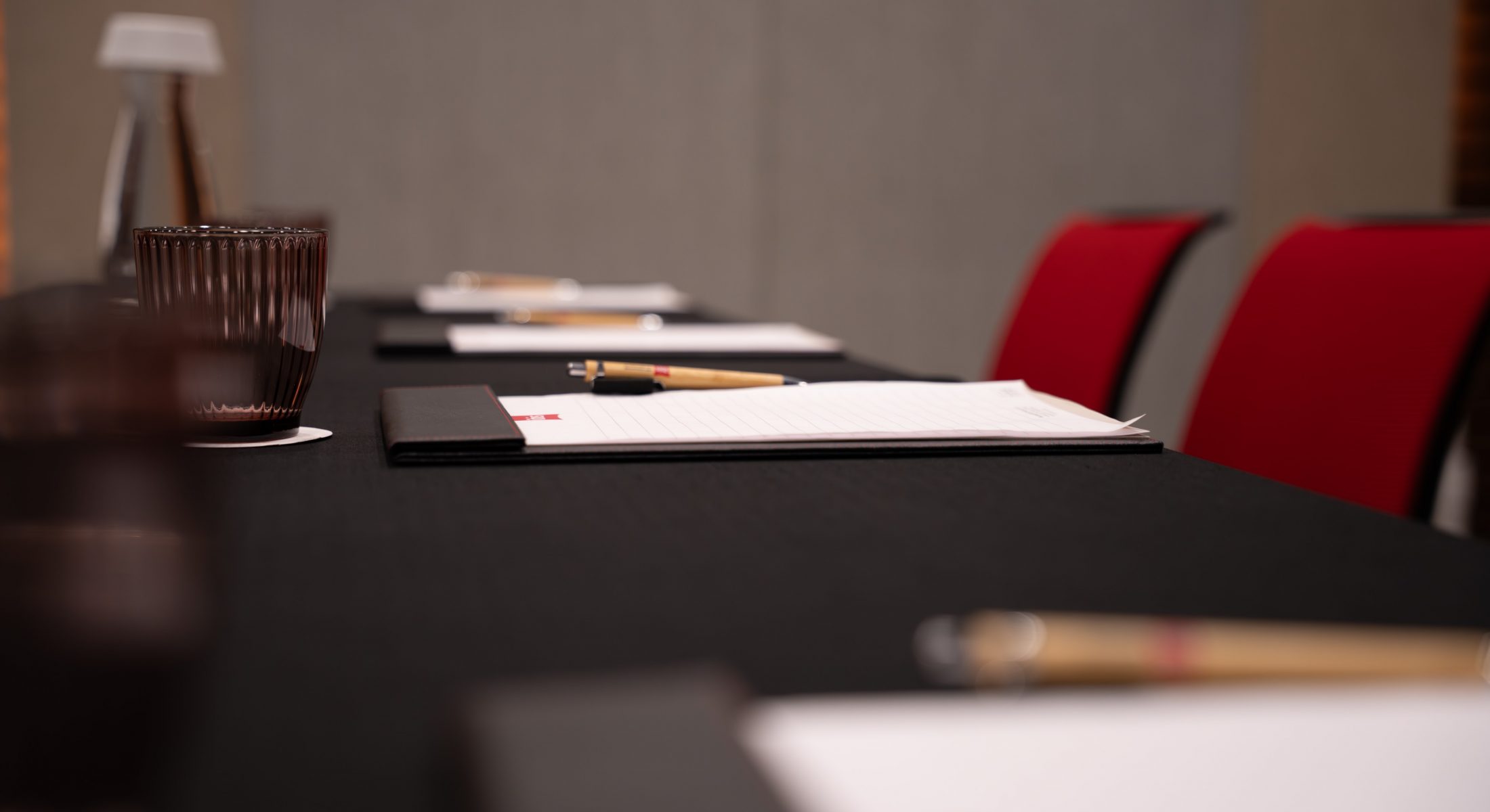 Close-up of writing pads, pens and drinking glasses on a black-covered meeting table, with red chairs behind.