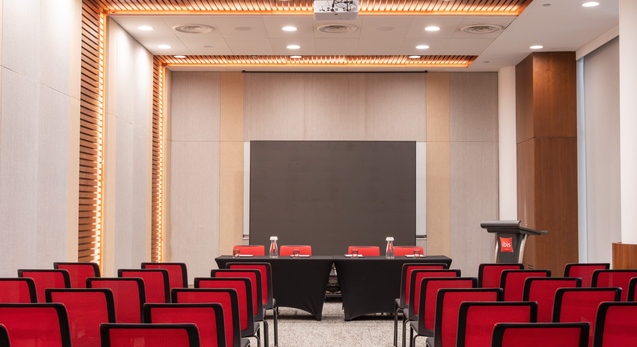 Central aisle between rows of red chairs leading to a four-seat panel table and lectern beneath a ceiling-mounted projector.