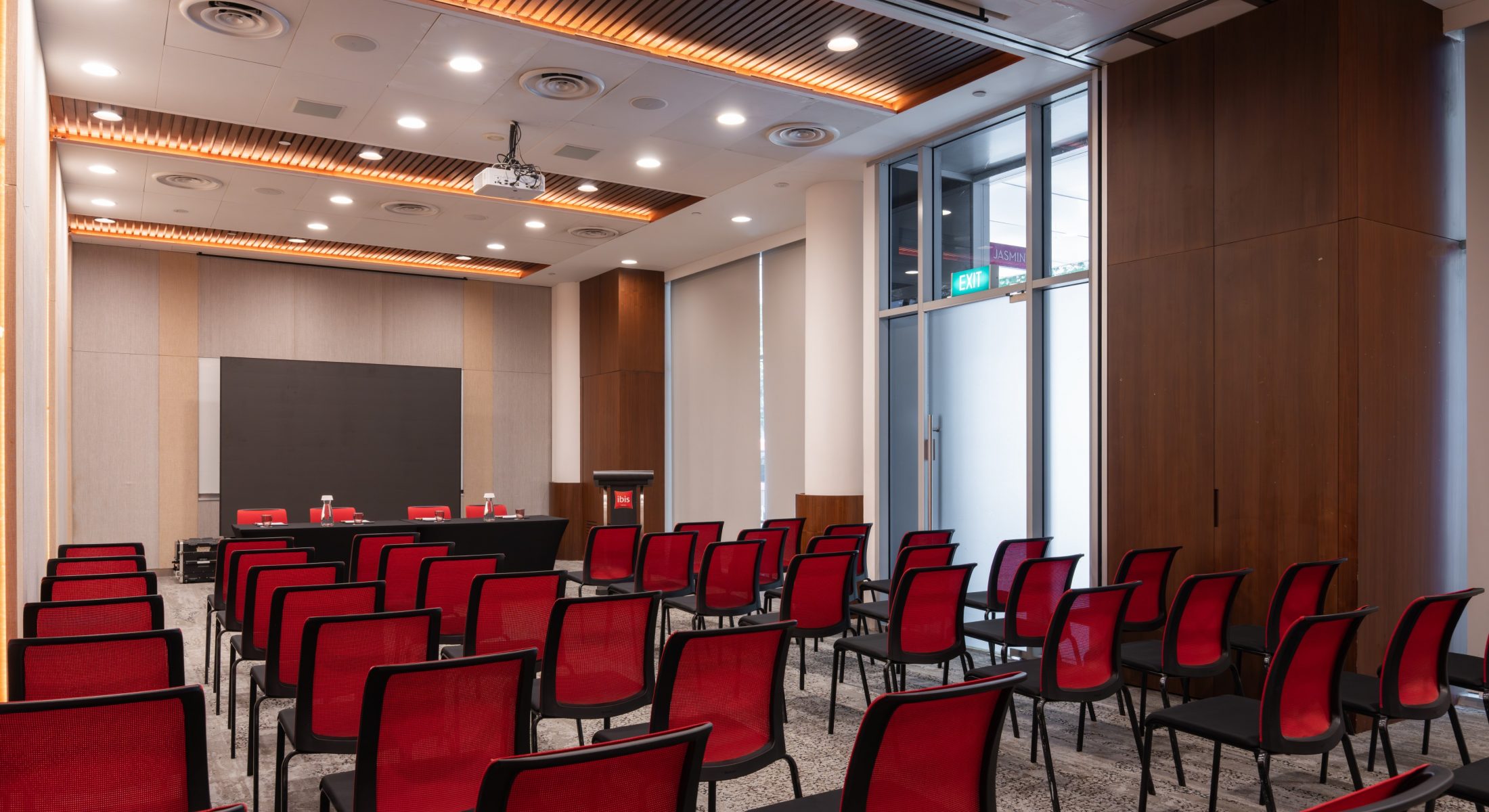 Wide view of theatre seating facing a panel table and lectern, with tall windows and a ceiling-mounted projector.