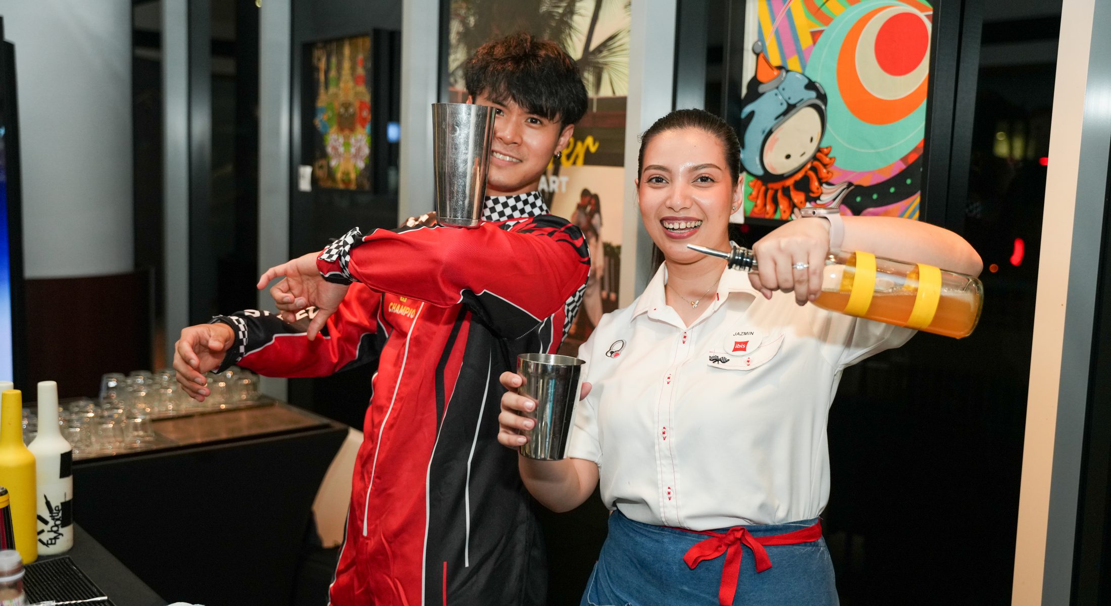 Two people demonstrating cocktail mixing with metal shakers and a bottle, with colourful artwork behind them.
